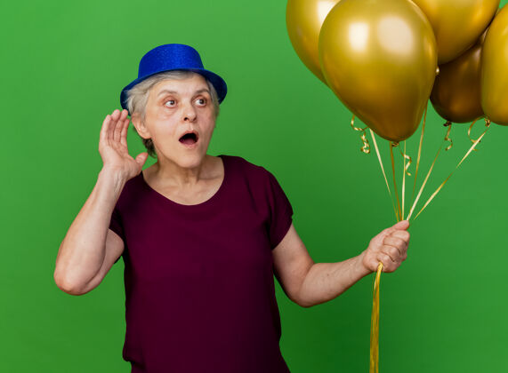女人震惊的老妇人戴着党的帽子举行氦气球保持手在耳朵后面看着绿色的一面气球耳朵手