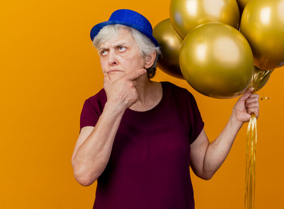 穿体贴的老妇人戴着聚会帽 把手放在下巴上 手里拿着氦气球 看着橙色的天空聚会起来手