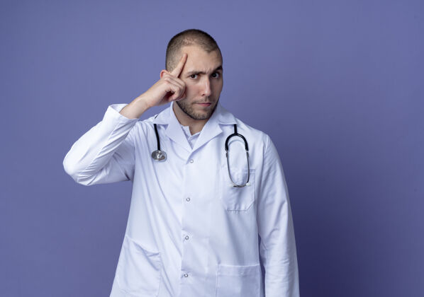 医学体贴的年轻男医生 穿着医用长袍 脖子上戴着听诊器 把手指放在隔离在紫色墙上的头上表情年轻听诊器