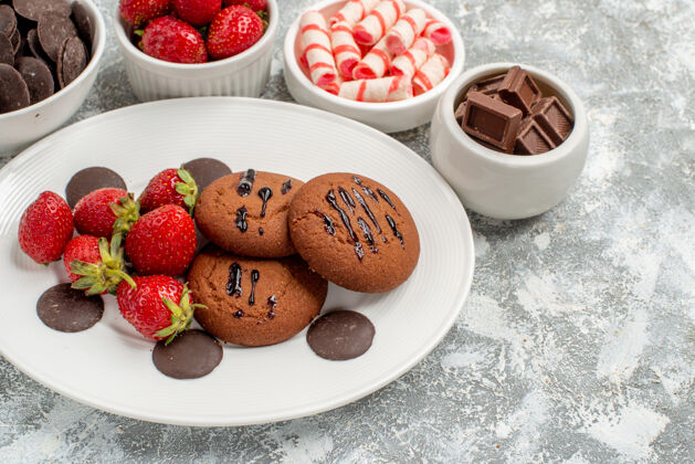 糖果下半部分是白色椭圆形盘子上的饼干草莓和圆形巧克力 灰白色桌子上的碗里放着糖果草莓和巧克力底部一半桌子