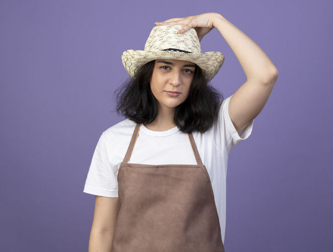 女人自信的年轻黑发女园丁身着制服 手戴紫墙上隔离的园艺帽自信女人花园