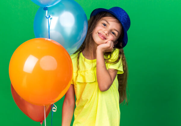 生日可爱的白人小女孩 戴着蓝色派对帽 手放在脸上 手里拿着氦气球 隔离在绿色墙壁上 留有复制空间小抱孤立