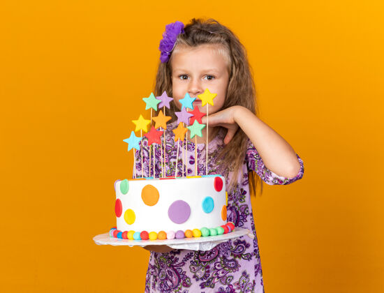 下巴高兴的金发小女孩拿着生日蛋糕 把手放在下巴上 隔离在橙色的墙上 留着复制空间抱着金发孤立