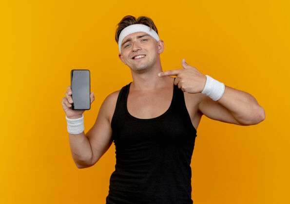 市民面带微笑的年轻英俊的运动型男子戴着头带和腕带展示和指着孤立在橙色墙上的手机感觉帅气衣服
