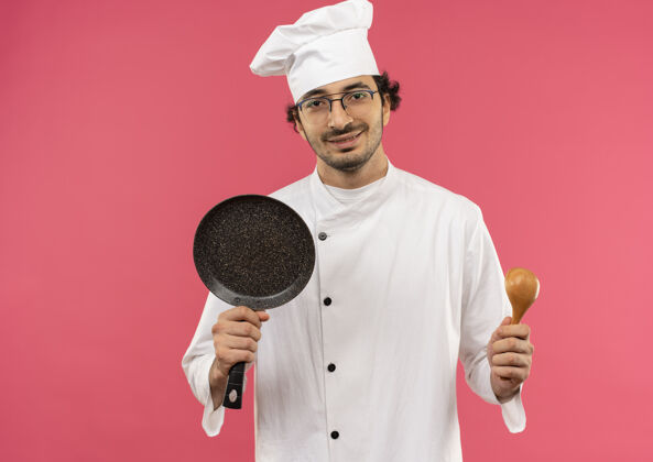 眼镜年轻的男厨师穿着厨师制服 戴着眼镜 手里拿着煎锅和勺子勺子烹饪请