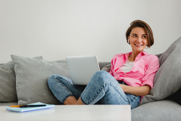 工人一个穿着粉色衬衫的微笑女人 在家里的沙发上轻松地坐在桌子旁 在家里用笔记本电脑上网笔记本电脑室内坐姿