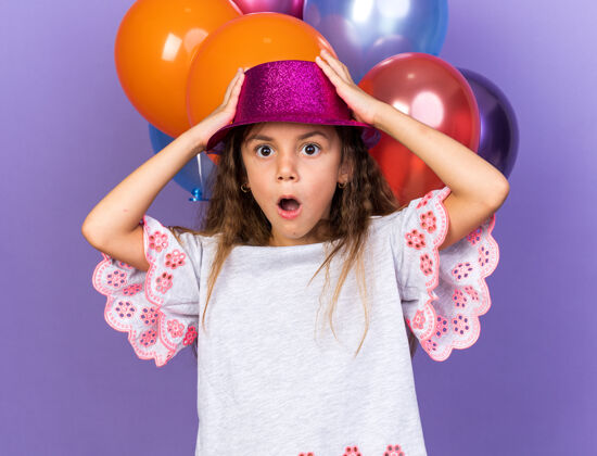 白种人震惊的小白种女孩 戴着紫色派对帽 手放在帽子上 站在氦气球前 在紫色的墙上 留着复制空间小孤立前面
