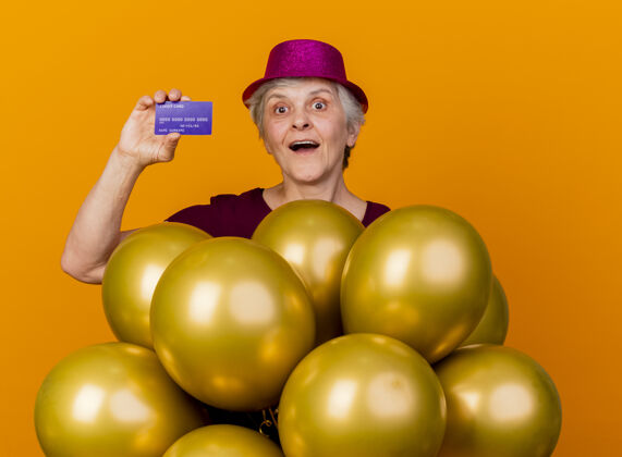 表情惊讶的老妇人戴着派对帽站在氦气球上 手里拿着信用卡 隔离在橙色的墙上举行人老人