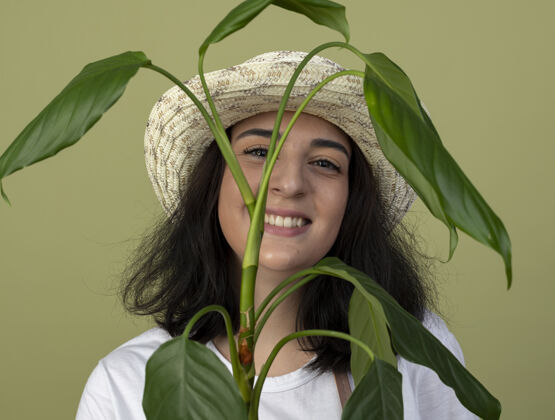 站微笑的年轻黑发女园丁穿着制服 戴着园艺帽 站在橄榄绿墙上孤立的植物后面花园表情帽子