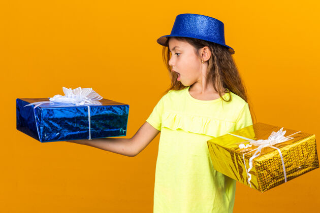 孤立惊讶的小白种人女孩拿着蓝色的聚会帽 看着孤立在橙色墙上的礼品盒和复印空间惊喜白种人盒子