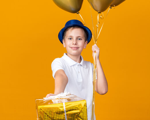 拿着高兴的年轻斯拉夫男孩与蓝色党帽子举行氦气球和礼品盒隔离在橙色墙上的复制空间男孩气球孤立的