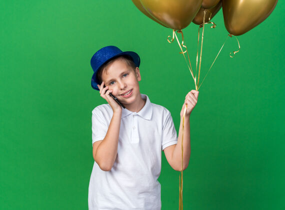 年轻的高兴的年轻斯拉夫男孩 戴着蓝色的派对帽 手里拿着氦气球 在绿色的墙壁上讲着电话 还有复印空间帽子氦孤立的