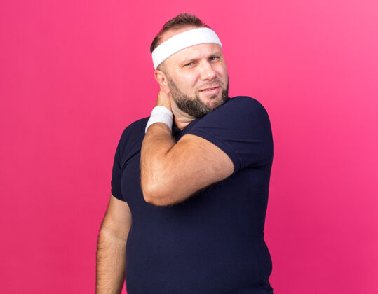 戴疼痛的成年斯拉夫运动男子戴着头带和腕带把他的手放在脖子上隔离在粉红色的墙壁与复制空间头带男人成人