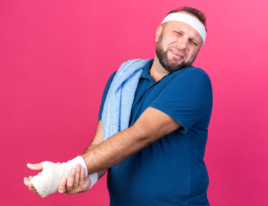 毛巾疼痛的成年斯拉夫运动型男子肩上带着毛巾戴着头带和腕带 手被隔离在粉色墙壁上 留有复制空间抱着戴运动