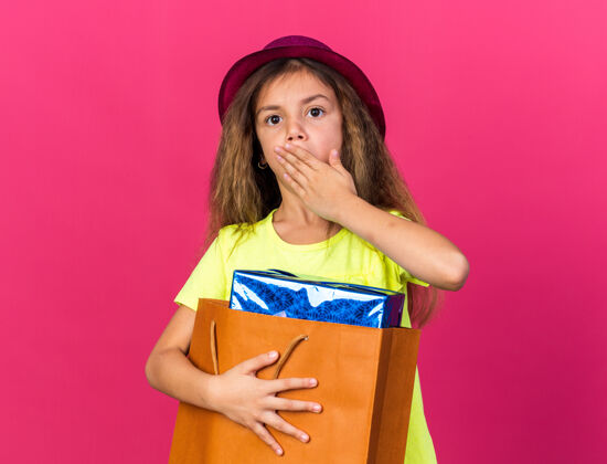 孤立一个惊讶的白人小女孩 戴着紫色派对帽 手放在嘴上 手里拿着一个装在纸袋里的礼品盒 隔离在粉红色的墙上 还有复印空间手拿着小