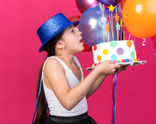 白种人惊讶的年轻白人女孩戴着蓝色的派对帽 看着生日蛋糕 手里拿着氦气球 隔离在粉红色的墙上 还有复制空间孤立拿着女孩