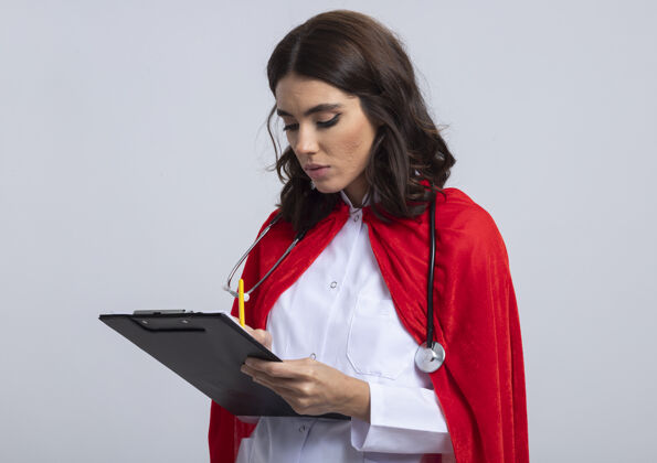 铅笔自信的女超人穿着医生制服 戴着红色斗篷和听诊器 用铅笔写在剪贴板上 隔离在白墙上人斗篷姿势