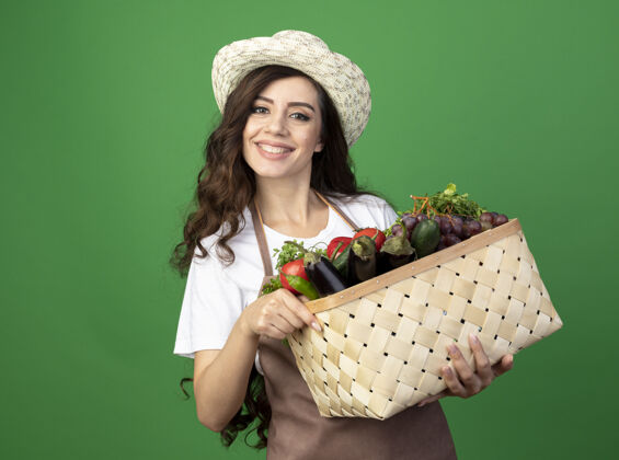 拿着身着制服 戴着园艺帽 面带微笑的年轻女园丁把菜篮子孤立地放在绿色的墙上人女人蔬菜