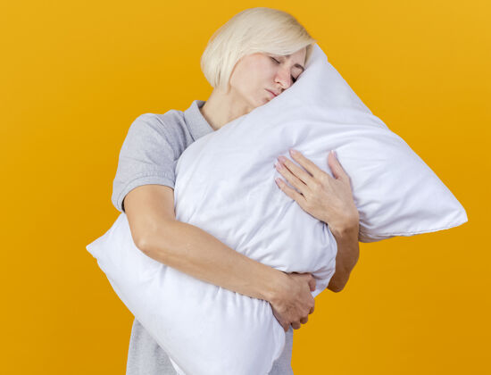 年轻昏昏欲睡的年轻金发病女人拥抱 把头放在隔离在橙色墙上的枕头上人表情市民