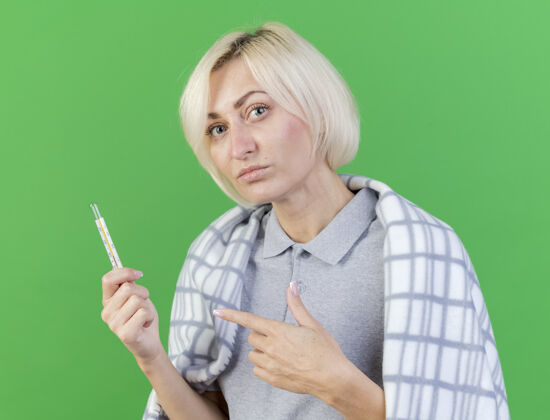 疾病年轻自信的金发病女人裹着格子呢的衣夹 指着隔离在绿色墙上的温度计体温计金发格子