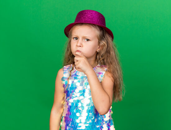 女孩体贴的金发小女孩 戴着紫色的派对帽 手放在下巴上 在绿色的墙壁上看起来很孤立 留着复印空间孤立金发手