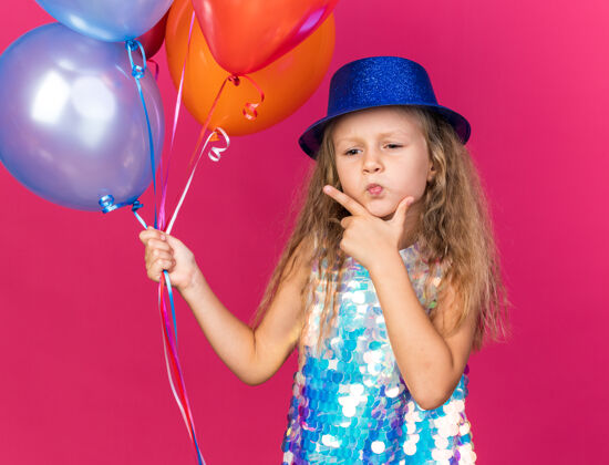 下巴体贴的金发小女孩 戴着蓝色派对帽 手放在下巴上 手里拿着氦气球 孤立地放在粉红色的墙上 还有复印空间思想抱派对