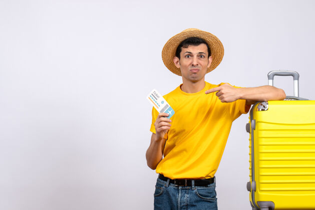 视图正面图穿着黄色t恤的年轻人站在黄色手提箱旁指着车票T恤指向黄色