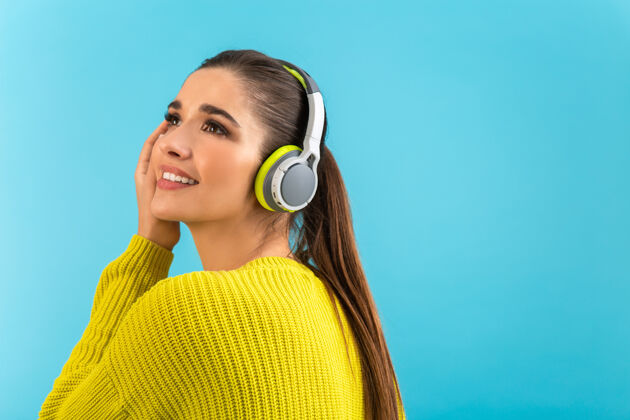 可爱戴着无线耳机听音乐的女人开心地穿着黄色针织毛衣摆着蓝色的姿势女人设备音频
