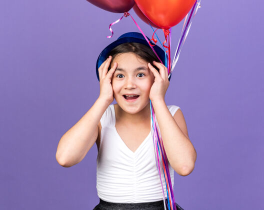 氦惊讶的年轻白人女孩戴着蓝色派对帽 手放在脸上 手里拿着氦气球 隔离在紫色的墙上 留着复制空间白种人气球手