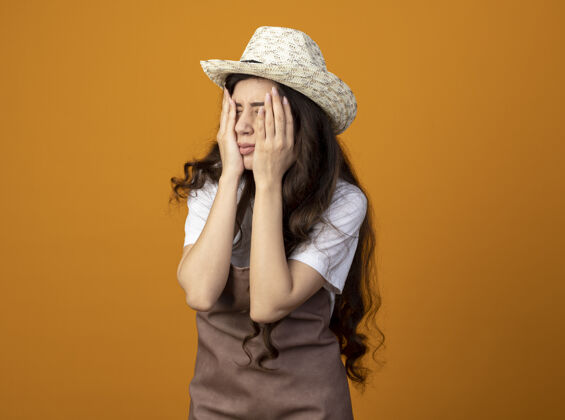 穿着失望的年轻女园丁穿着制服 戴着园艺帽 把手放在隔离在橙色墙上的脸上失望人表情