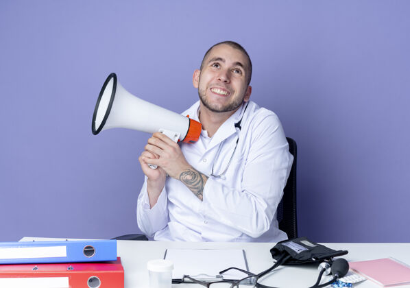 人面带微笑的年轻男医生 穿着医用长袍 手持听诊器 坐在办公桌旁 手里拿着工作工具 举着扬声器 孤零零地看着紫色的墙上桌子医生紫色