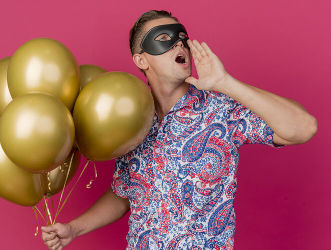 侧线年轻的派对小伙儿戴着化装眼罩看着身边拿着气球 喊着有人被隔离在粉红色粉色小伙子派对