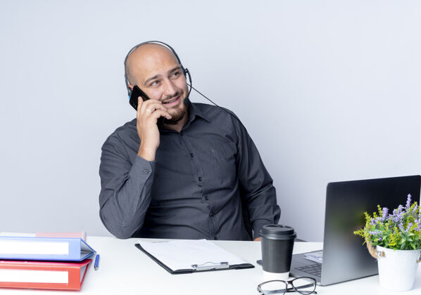 工具微笑着的年轻秃头呼叫中心男子戴着耳机坐在办公桌旁 工作工具在一旁看着 隔着白墙上的电话交谈姿势电话人