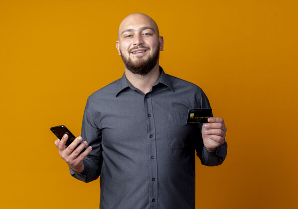 秃头微笑的年轻秃头呼叫中心男子拿着信用卡和手机孤立在橙色的墙上电话抱着电话