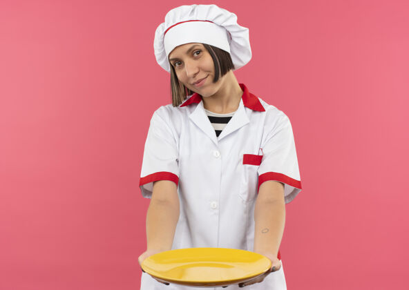 人身着厨师制服的年轻女厨师高兴地把盘子朝前伸出 孤立地放在粉红色的墙上表情衣服烹饪