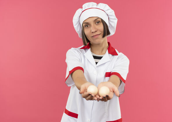 朝向身着厨师制服的年轻女厨师高兴地把鸡蛋伸向隔离在粉红色墙上的前面人感情姿势