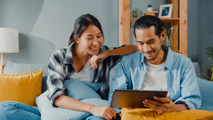 男人快乐的亚洲年轻漂亮的夫妇男女坐在沙发上用平板电脑在新房子里在线购买家具夫妇沙发发现