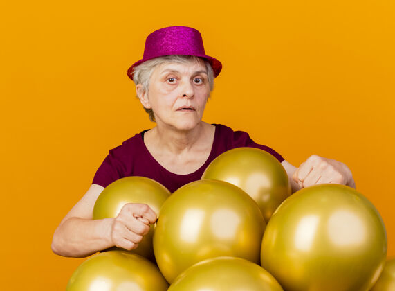 手自信的老妇人戴着派对帽站在那里 手里拿着氦气球 指着隔离在橙色墙上的浴室手势表情氦姿势