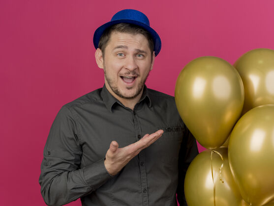 欢乐快乐的年轻人戴着蓝色的帽子拿着聚会 用手指着粉红色的气球举行气球穿