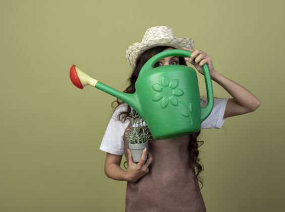 人焦急的年轻女园丁身穿制服 戴着园艺帽 手持花盆 透过橄榄绿墙上的浇水罐望着前方姿势脸市民