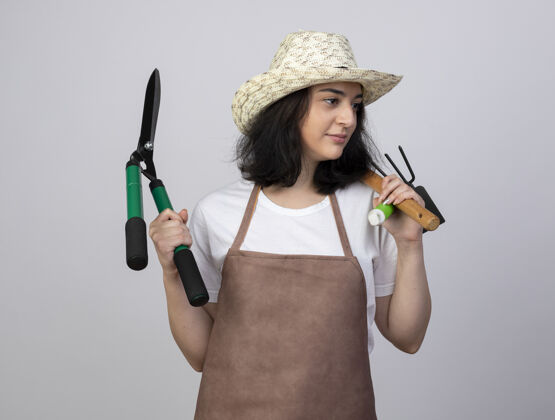 表情高兴的年轻黑发女园丁穿着制服戴着园艺帽手持园艺工具看着隔离在白色墙上的一面制服拿着人