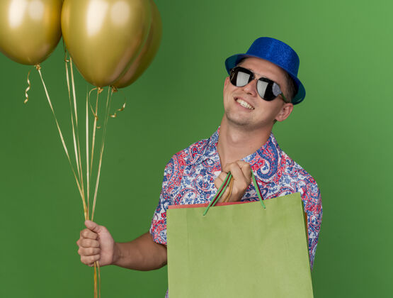 拿着微笑的年轻人戴着蓝色帽子 戴着眼镜 手里拿着礼品袋 气球孤立在绿色上年轻人气球小伙子
