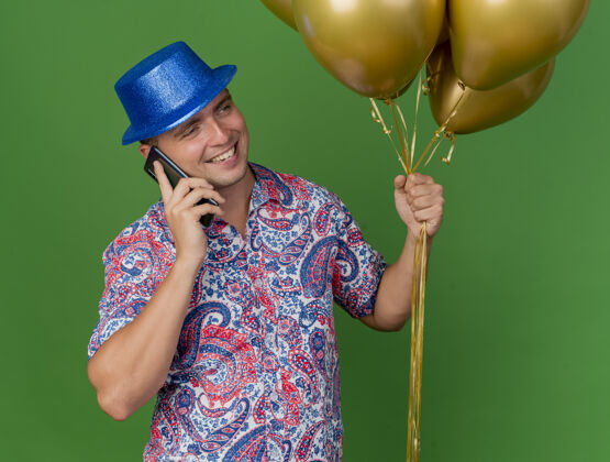 气球带着微笑的年轻人戴着蓝色的帽子拿着气球在电话里说绿色的隔离派对小伙子蓝色