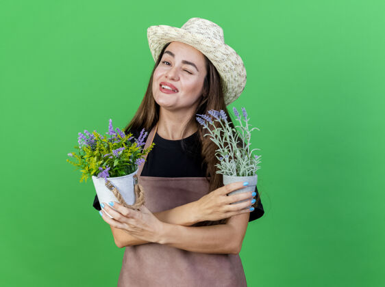 花盆闪烁着微笑的美丽园丁女孩穿着制服戴着园艺帽拿着和交叉的花盆隔离在绿色交叉花帽子