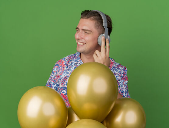 耳机高兴的年轻人穿着五颜六色的衬衫 戴着耳机 站在绿色的气球后面看派对站派对拜托