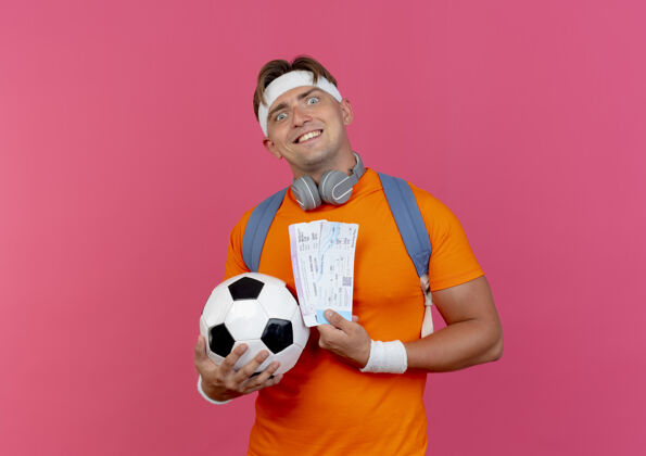 穿着面带微笑的年轻英俊的运动型男子戴着头带和腕带 脖子上背着一个戴着耳机的包 手里拿着飞机票 粉色墙上隔着一个足球背脖子衣服