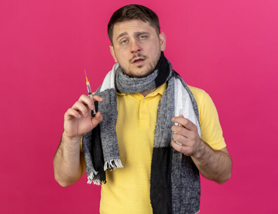 疾病自信的年轻金发病夫戴着围巾拿着注射器和安瓿隔离在粉红色的墙上姿势年轻人男人