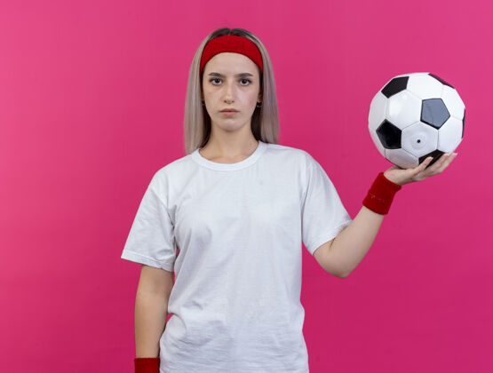 女人自信的年轻运动女性戴着背带 戴着头带和腕带 把球隔离在粉红色的墙上腕带年轻人