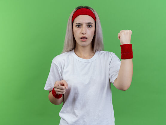女人笨手笨脚的年轻运动型女子戴着护腕 戴着头带和腕带 把拳头和手指放在隔离的绿色墙上穿无知拳头