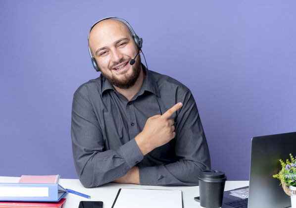 耳机微笑着的年轻秃头呼叫中心男子戴着耳机坐在办公桌旁 工作工具指着隔离在紫色墙上的一侧市民电话感觉